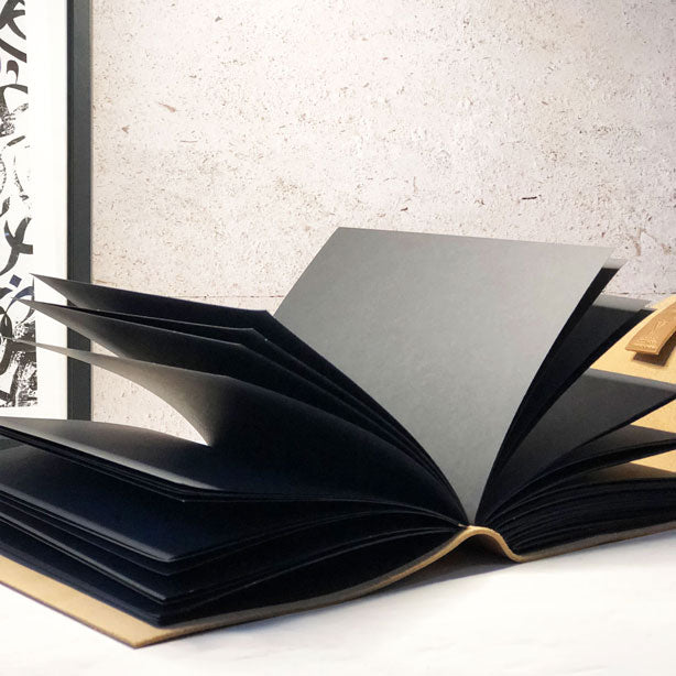 A large leather journal with black paper pages, partially open, displaying its thick pages and binding. The journal is on a light-colored surface with a textured wall in the background.