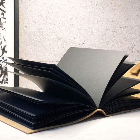 A large leather journal with black paper pages, partially open, displaying its thick pages and binding. The journal is on a light-colored surface with a textured wall in the background.
