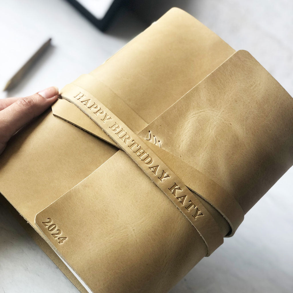 Tan leather-bound notebook with 'Happy Birthday Katy' and '2024' embossed on it, held by a hand.